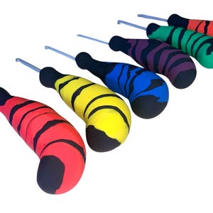 May include: A set of six crochet hooks with ergonomic, thick handles. Each handle features a distinct colour—red, yellow, blue, purple, green, and orange—with black tiger-stripe patterns. The metal hooks are silver and extend from the top of each handle.