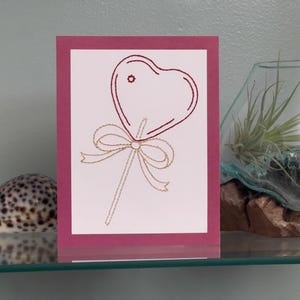 May include: A greeting card with a heart-shaped lollipop design. The heart and stick are outlined in red thread, with a gold bow at the base. The card is set against a pink background.