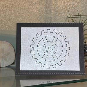 May include: A framed card with a gear design and the letters "VS" in the center. The card has a white background with a black gear outline and a dark gray border. The card is displayed on a glass shelf.