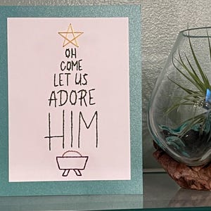 May include: A Christmas card with the words "Oh Come Let Us Adore Him" in black script. The card has a gold star and a manger illustration. The card is on a teal background.