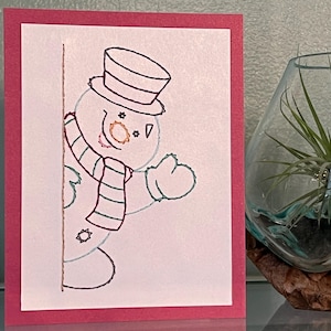 Pode incluir: Um cartão de felicitações feito à mão com um design de boneco de neve caprichoso. O boneco de neve, delineado a preto, usa um chapéu alto e um cachecol listrado. O cartão tem um interior branco com uma borda rosa, perfeito para as festas de inverno.