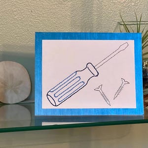 May include: A framed artwork featuring a blue-handled screwdriver and two screws, all in a simple line drawing style. The artwork has a blue border and is displayed on a glass shelf.