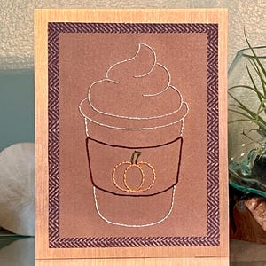 May include: A fall-themed greeting card featuring an embroidered coffee cup with whipped cream and a pumpkin. The card has a brown background with a dark purple border and a wooden frame. The pumpkin is embroidered in gold thread.