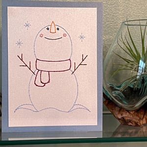 May include: A handmade greeting card with a whimsical snowman design. The snowman is drawn with blue outlines and features a carrot nose, twig arms, and a red scarf. Snowflakes are scattered around the snowman on a white background with a light blue border.
