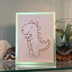 May include: A greeting card featuring a hand-drawn dinosaur illustration with red accents. The dinosaur is white with red spines and details. The card has a gold border and is displayed with a glass terrarium and decorative shells.