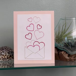 May include: A greeting card with a pink border features an embroidered design of hearts emerging from an envelope. The hearts are in shades of pink and red. A decorative glass terrarium and a seashell are also visible.