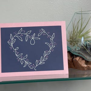May include: A greeting card with a navy blue center and a pink border. A white floral heart design is centered on the navy blue background. The card is displayed on a glass shelf with a decorative terrarium.