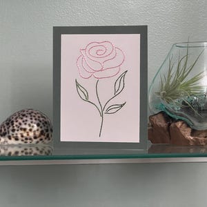 May include: A framed artwork with a pink rose embroidery on a white background, framed by a grey border. A seashell and a glass terrarium with a plant are also on a glass shelf.