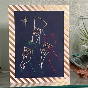 May include: A navy blue greeting card with an embroidered design of three kings and a star. The card is framed with a white and gold striped border. A decorative glass vase with a plant is in the background.