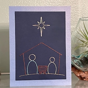 May include: A handmade greeting card featuring an embroidered nativity scene. The card has a dark blue background with a gold star and a brown and gold embroidered stable with two figures. The card is framed with a light blue border.