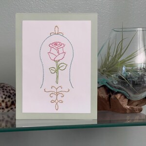 May include: A framed card with a hand-stitched design of a pink rose inside a bell jar. The card is set against a light green background. A glass terrarium with a plant and a seashell are also visible.