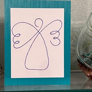 May include: A greeting card featuring a minimalist angel design. The angel is drawn with a single, continuous dark blue line on a white background. The card is set within a teal blue wooden frame, creating a simple yet elegant aesthetic.