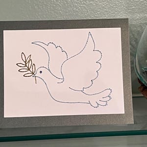 May include: A framed artwork featuring a white dove carrying an olive branch, outlined in blue stitching. The dove is centered on a white background, set within a gray frame. The artwork is displayed on a glass shelf.