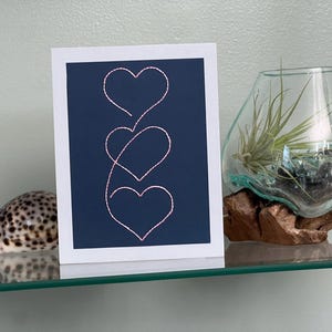 May include: A greeting card featuring three linked pink hearts against a navy blue backdrop. The card is on a glass shelf, alongside a glass terrarium with a plant and a seashell.