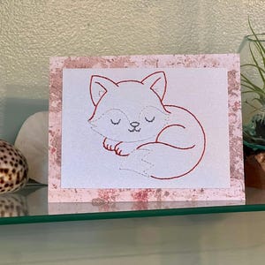 May include: A greeting card featuring a red outline drawing of a sleeping fox. The fox is curled up on a white background, framed by a pink and white patterned border. The card is displayed on a glass shelf.