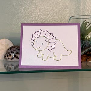 May include: A greeting card with a purple border features a gold-outlined illustration of a triceratops dinosaur on a white background. The card is displayed on a glass shelf with decorative items, including a seashell and a glass terrarium.