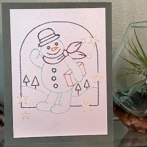 May include: A handmade greeting card featuring a stitched snowman design. The snowman wears a hat and scarf, holding a gift. Gold stars and trees decorate the scene, set against a white background with a gray border. A festive winter theme.