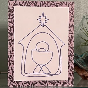 May include: A Christmas card featuring a hand-drawn nativity scene with a star above the stable. The card has a white center with a purple and white floral border.