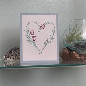 May include: A greeting card with a heart-shaped floral embroidery design. The heart is outlined in green thread and features three red tulips. The card is displayed on a glass shelf with a decorative glass terrarium and a speckled stone.