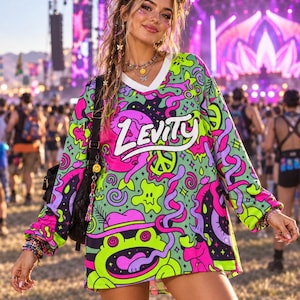 Puede incluir: Una camiseta de manga larga con cuello en V que presenta un estampado gráfico vibrante y multicolor con formas abstractas en verde neón, rosa intenso y morado, estrellas, un símbolo de la paz y una rana. La palabra Levity está impresa en blanco.