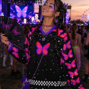 May include: A black long-sleeve jersey featuring a vibrant pink and orange butterfly pattern, a white and black checkered hem, and a small Cosmic Disco label. The jersey is paired with a black folding hand fan decorated with matching butterflies.