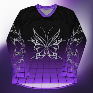 May include: A long-sleeved jersey in black and purple, with a V-neck. It showcases a large butterfly design and silver patterns on the sleeves. The background has a grid pattern that fades from purple to black. The jersey is a striking piece.