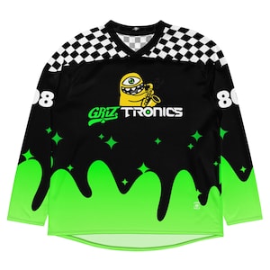 May include: Black long-sleeve jersey with a neon green slime design at the bottom. The jersey features a cartoon character playing a saxophone, the text "Griztronics", and the numbers "808" on the sleeves. The shoulders have a black and white checkered pattern.