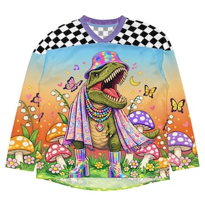 Puede incluir: Camiseta de manga larga con un dinosaurio de dibujos animados que lleva un sombrero, una bufanda y botas de arcoíris, rodeado de setas y mariposas. La camiseta tiene un estampado de cuadros en los hombros y un cuello en V. El fondo hace una transición de azul a naranja.
