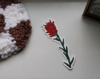 May include: A red and green flower sticker with a white background. The flower has a red bloom with green leaves and a stem.