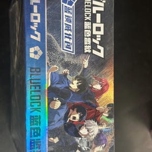 Puede incluir: Una caja azul y negra con personajes de anime en poses din&aacute;micas. La caja muestra texto japon&eacute;s y el t&iacute;tulo "BLUELOCK" en blanco. Tambi&eacute;n se ve el texto "BLUELOCK 蓝色监狱" y "蓝锁威化包".