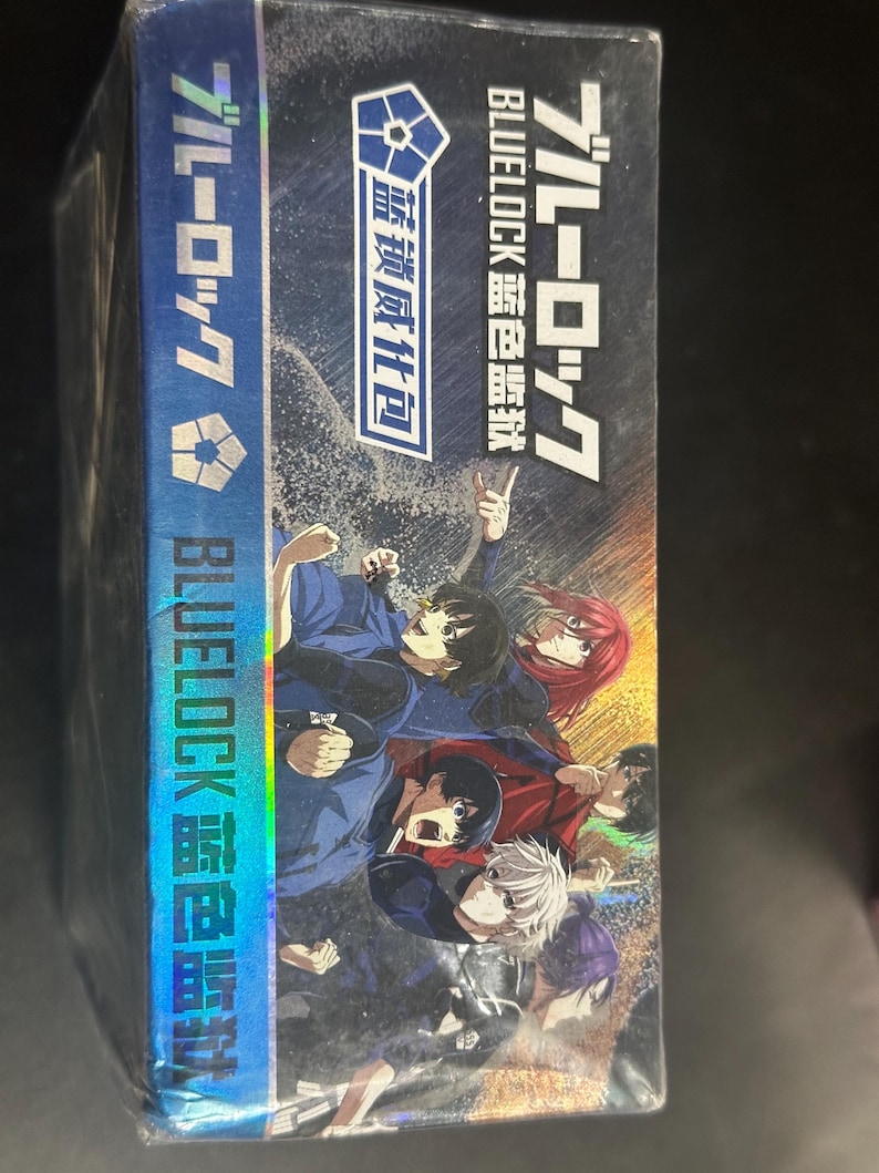Puede incluir: Una caja azul y negra con personajes de anime en poses din&aacute;micas. La caja muestra texto japon&eacute;s y el t&iacute;tulo "BLUELOCK" en blanco. Tambi&eacute;n se ve el texto "BLUELOCK 蓝色监狱" y "蓝锁威化包".
