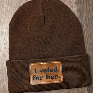 May include: A brown knit beanie with a rectangular leather patch. The patch features the text "I voted for her." in a serif font. The beanie is made of a soft, ribbed knit material.
