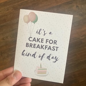 May include: A white card with the text "it's a CAKE FOR BREAKFAST kind of day" and a cake illustration. The card features three balloons in pastel colors and small confetti-like sprinkles.