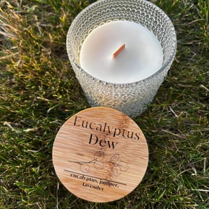 May include: A white wax candle in a clear glass jar with a textured hobnail pattern and a wooden wick. The round bamboo lid features the text Eucalyptus Dew, a line drawing of a eucalyptus branch, and the words eucalyptus, juniper, lavender.