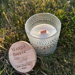 May include: A clear glass candle holder with a textured hobnail pattern containing white wax and a wooden wick. Beside it lies a round bamboo lid engraved with the text Mango Breeze, a tree illustration, and the words peony, sandalwood, mango.