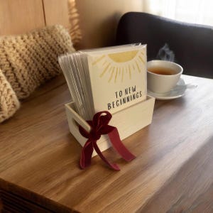 May include: A white wooden box with a red ribbon holds a stack of greeting cards with a yellow sun and the text "To New Beginnings".
