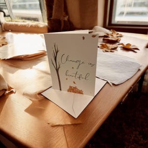 May include: A white card with the text "Change is beautiful" written in black cursive. The card has a brown branch and a pile of brown leaves in the background.