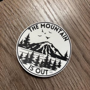 May include: A circular white sticker with a black mountain and forest design. The text "THE MOUNTAIN IS OUT" is printed around the edge. Two birds are flying above the mountain.