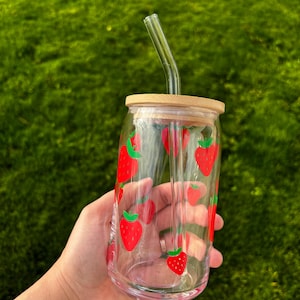 May include: A clear glass tumbler featuring a repeating pattern of red strawberries with green leaves. The glass includes a light-colored bamboo lid with a circular opening and a matching clear glass straw that is bent at the top for drinking.