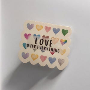 May include: A white sticker with a scalloped edge features a colorful array of watercolor hearts. The sticker has the text "LOVE OVER EVERYTHING" in black lettering.