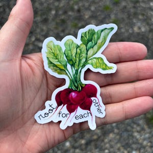 May include: A sticker featuring a watercolor illustration of radishes with green leaves and the text "root for each other". The radishes are a deep red color.