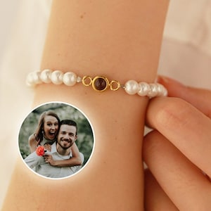May include: A white pearl bracelet with a gold charm featuring a photo of a couple. The charm is a round, clear glass cabochon.