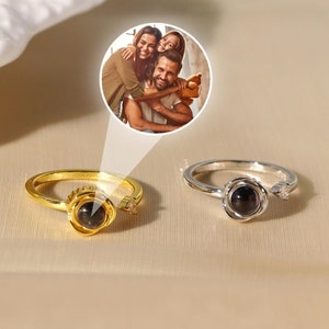 May include: Two silver rings with a black rose design. The rings have a small clear stone on the side. The rings are on a beige surface. One ring is gold and the other is silver.
