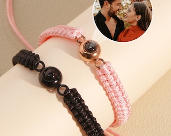 Könnte beinhalten: Zwei Freundschaftsarmbänder mit schwarzen und rosafarbenen Schnüren. Jedes Armband hat einen kleinen runden Anhänger mit einem versteckten Bild darin. Das Bild zeigt ein Paar, das sich anschaut.