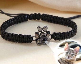 May include: Black braided bracelet with a black metal paw print charm. The charm has a small clear glass insert that shows a picture of a cat.