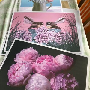 May include: Three framed prints of floral arrangements. The top print features a bouquet of pink, white, and blue flowers in a brown ceramic vase. The middle print features two black and white birds perched on a cluster of pink hydrangeas. The bottom print features a cluster of five pink peonies on a black background.