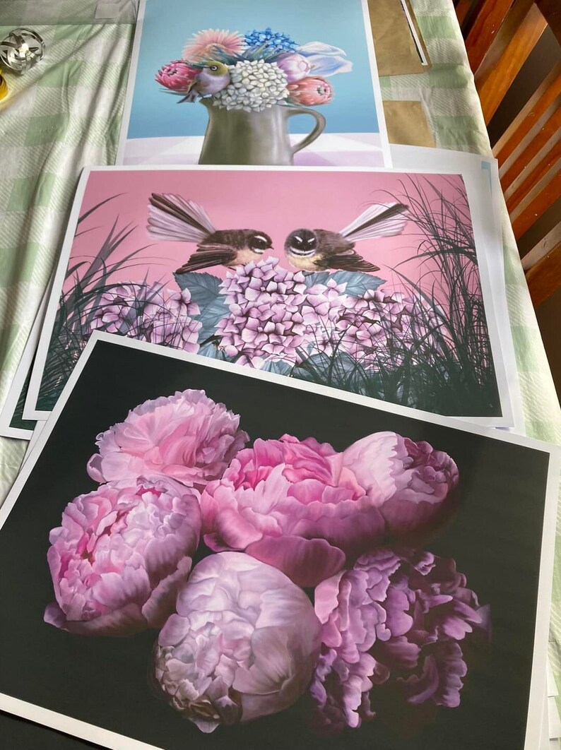 May include: Three framed prints of floral arrangements. The top print features a bouquet of pink, white, and blue flowers in a brown ceramic vase. The middle print features two black and white birds perched on a cluster of pink hydrangeas. The bottom print features a cluster of five pink peonies on a black background.
