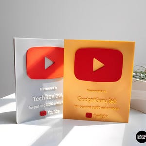 Puede incluir: Dos premios YouTube Play Button. Uno es plateado con un botón de reproducción rojo, el otro es dorado con un botón de reproducción rojo. El premio plateado es para TechReview, y el premio dorado es para Gadget Guru 360. Ambos premios tienen el logo de YouTube.