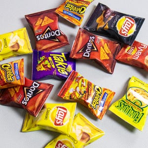 May include: A collection of various snack-sized chip bags, including Doritos, Cheetos, Lays, and Takis, in a variety of colors. The bags are scattered on a light gray surface, showcasing the different brands and flavors.