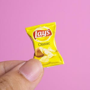May include: A miniature yellow Lay's Classic potato chip bag with the brand's logo and the word "Classic" printed on it. The bag contains images of potato chips. The bag is held against a pink background.
